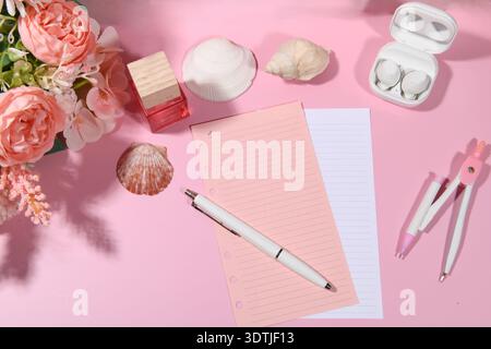 Feminines pinkfarbenes Display mit Kunstblumen, Muscheln, airpods und Schreibwaren, Kompass, Stift und Parfüm. Hohe Winkelansicht auf rosafarbener Oberfläche. Stockfoto