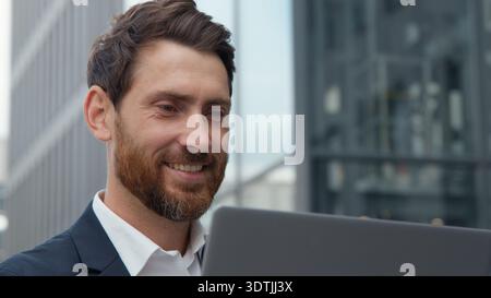 Close-up lächelnder erfolgreicher kaukasischer reifer Geschäftsmann, der an Laptop im Stadtporträt arbeitet, Geschäftsmann mittleren Alters männlicher freudiger Chat Stockfoto