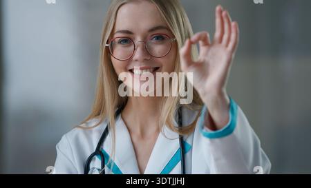 Kaukasische Ärztin lächelt zeigt OK Geste glückliche Krankenschwester weibliches Mädchen Lächeln in die Kamera zeigt OK Hand in der Klinik erfolgreich Stockfoto