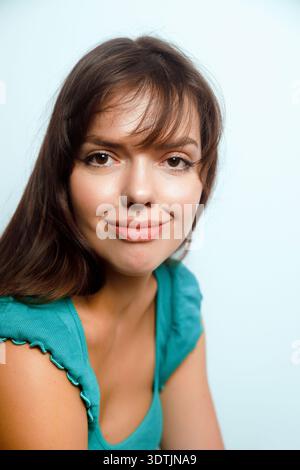 Eine Frau mit langen dunklen Haaren lächelt, während sie ein blaues Oberteil trägt. Sie sitzt vor hellem Hintergrund in einem Studio. Die Umgebung ist hell, und sie Stockfoto