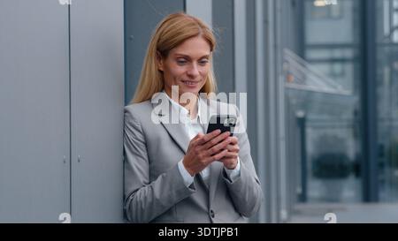 Lächelnde kaukasische Geschäftsfrau mittleren Alters, die ein Mobiltelefon hält, die Wand des Büros, das Firmengebäude des Unternehmens, das Lächeln der positiven 40er Jahre Stockfoto