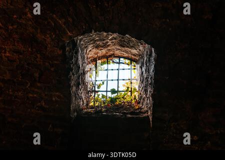 Historisches Kerkerfenster der Festung mit Eisenstangen und starkem Licht dahinter, das Gefangenschaft, Isolation, Flucht und Kontrast zwischen Dunkelheit und repräsentiert Stockfoto