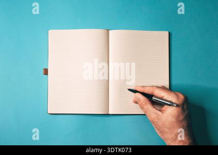 Overhead-Aufnahme eines offenen Hardcover-Notizbuchs mit linierten Seiten und schwarzem Stift, der über leerem Blatt auf blauem Hintergrund gehalten wird, minimalistisches Schreibkonzept. Auswahl Stockfoto