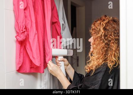 Eine rothaarige Frau benutzt ein Dampfbügeleisen, um zu Hause ein rosafarbenes Hemd zu drücken. Die Nahaufnahme erfasst ihre Konzentration und die Effizienz des Dampfbügeleisens in cl Stockfoto