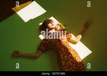 Eine Frau in einem Blumenkleid liegt friedlich auf einer grünen Oberfläche, in warmes Sonnenlicht getaucht. Ihre entspannte Pose und ihr heiterer Ausdruck sorgen für eine ruhige und ruhige Atmosphäre Stockfoto