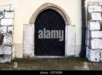 Ein Vollformatblick auf eine alte schwarze mittelalterliche Tür mit einem markanten Bogenrahmen aus Stein vor einer hellen cremefarbenen Wand. Die Tür selbst besteht aus da Stockfoto