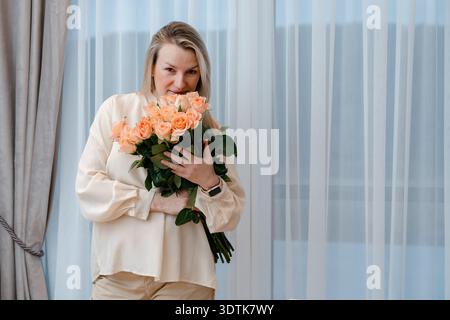 Blonde Frau, die Blumen in der Nähe der Brust hält. Ideal für Valentinstag, Geburtstag oder Danksagungskampagnen. Gemütliches familiäres Ambiente. Stockfoto