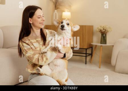 Lächelnde Frau mit entzückendem Corgi mit humorvollem Gesichtsausdruck im gemütlichen Wohnzimmer. Das Konzept des Besitzes von Haustieren, der Begleitung und des fröhlichen Wohnens Stockfoto