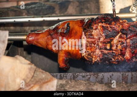 Ein Nahaufnahme-Bild zeigt, wie ein ganzes Schwein langsam auf einer Metallspieße über einem offenen Feuer gebraten wird. Das Schwein hat ein herrlich goldbraunes und knuspriges s Stockfoto
