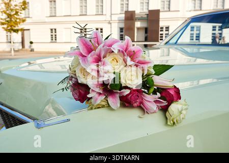 Ein wunderschöner Blumenstrauß aus rosa Lilien, weißen Rosen und dunkelrosa Rosen liegt auf der Motorhaube eines Vintage, hellgrünen Autos. Die Blumen sind geordnet Stockfoto