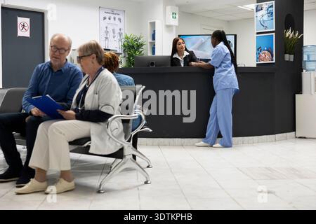 Älterer Arzt mit Klemmbrett, der die medizinischen Ergebnisse mit einem alten männlichen Patienten erklärt, der auf den Bänken der Krankenhauslobby sitzt. Asiatische Rezeptionistin und afroamerikanische Krankenschwester diskutieren an der Rezeption der Klinik. Stockfoto