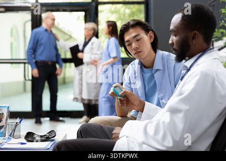 Der afroamerikanische Arzt misst den Blutzuckerspiegel während des Klinikbesuchs genau. Asiatischer Mann erhält aufmerksame Betreuung mit medizinischen Geräten, unterstützt Diagnose, Gesundheitsüberwachung und Behandlung Stockfoto