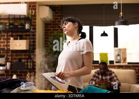 Die erschöpfte kaukasische Hausfrau bügelt Kleidung im Wohnzimmer, während der Ehemann mit dem Smartphone auf dem Sofa entspannt. Junge Frau, die alle Aufgaben alleine erledigt, müde ist und Rückenschmerzen hat. Stockfoto