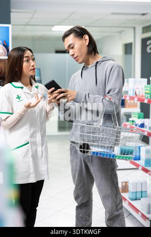 Der männliche Kunde liest mit dem Handy detaillierte Anweisungen zu Medikamenten, während er sich an den Apotheker wendet. Asiatischer Mann bittet Apothekerin um Hilfe bei der Auswahl des richtigen Produkts in der Apotheke. Stockfoto