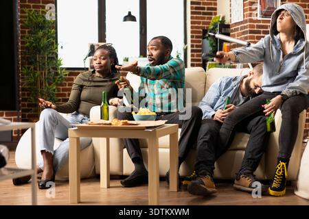 Multiethnische Gruppe im Wohnzimmer, die ein Live-Fußballspiel ansieht, enttäuscht, nachdem das Team das entscheidende Tor verpasst hat. Afroamerikanische und kaukasische Freunde runzeln im Fernsehen und reagieren frustriert nach dem Spielverlust. Stockfoto