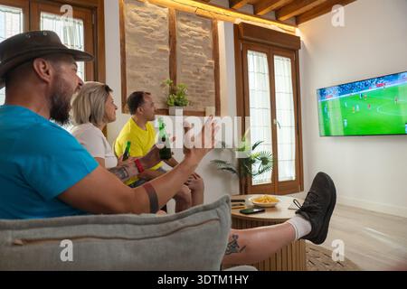 Eine Gruppe von Freunden, die nach einem Tor aufgeregt anfeuern, während sie ein Fußballspiel zu Hause im Fernsehen anschauen, Spaß, Freundschaft und Wochenendsportzeit genießen Stockfoto