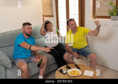 Eine Gruppe von Freunden jubelt und feiert ein Tor, während sie zu Hause ein Fußballspiel im Fernsehen anschauen. Aufgeregte Reaktionen, erhobene Arme, Bier, Snacks und Freunde Stockfoto