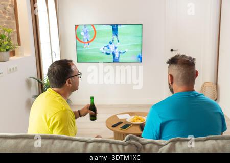 Zwei Freunde, die sich ein Fußballspiel von hinten im Fernsehen ansehen, während sie zu Hause auf einem Sofa sitzen. Ungezwungene Freizeit, sportliche Unterhaltung und entspannte Atmosphäre Stockfoto