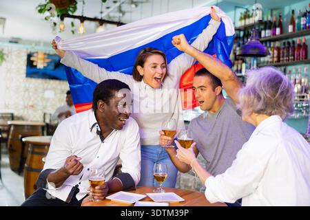 Fröhliche, vielfältige Fußballfans mit der russischen Flagge verbringen Zeit zusammen mit einem Pint Bier in der Kneipe Stockfoto