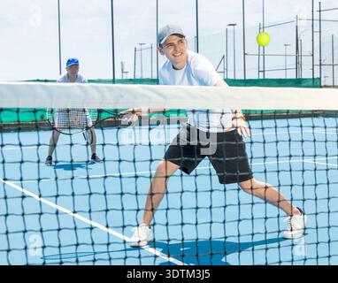 Porträt eines jungen Mannes, der im Sommer auf dem offenen Platz Tennis spielt, und Schläger schwingt, um den Ball über das Netz zurückzubringen. Sportler bereit für Volley Stockfoto