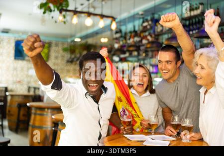 Begeisterte, multikulturelle Fußballfans feiern den Treffer ihrer Mannschaft im Pub mit erhobenen Händen Stockfoto