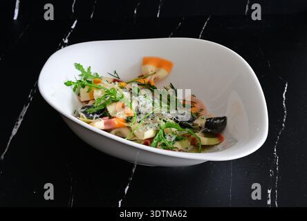 Gekochte bunte Pasta (Farfalle) mit Parmesankäse, Rucola, Dill und Fleischbällchen in einer weißen Schüssel vor schwarzem Hintergrund Stockfoto