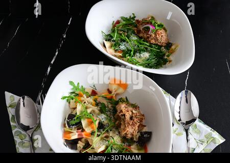 Gekochte bunte Pasta (Farfalle) mit Parmesankäse, Rucola, Dill und Fleischbällchen in einer weißen Schüssel vor schwarzem Hintergrund Stockfoto