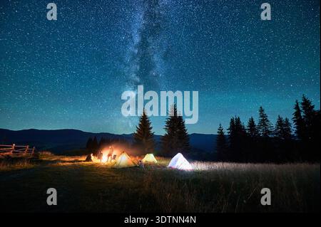 Silhouetten von Touristen, die auf dem Campingplatz am Lagerfeuer ruhen, mit leuchtenden Zelten in der Nähe unter Sternenhimmel. Die Milchstraße erstreckt sich über die Berge und wirft ein heiteres und magisches Licht über die beschauliche Berglandschaft. Stockfoto