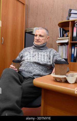 Senior-Mann sitzt in einem Büro. Zwei Kaffeetassen stehen neben ihm auf dem Schreibtisch Stockfoto