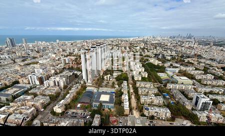 Aus der Vogelperspektive auf eine dichte Küstenstadt mit modernen Wolkenkratzern, Wohnblocks und einer Sportarena, die sich zum blauen Meer hin erstreckt. Stockfoto