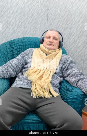 Ein älterer Mann entspannt sich in einem Stuhl mit Kopfhörern und genießt Musik in einem gemütlichen Innenbereich. Stockfoto