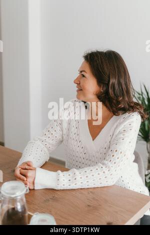 Frau in einem gemütlichen weißen Pullover sitzt an einem Holztisch und lächelt leicht, während sie wegschaut. Der schlichte Hintergrund und die dezente Einrichtung sorgen für eine entspannte Atmosphäre Stockfoto