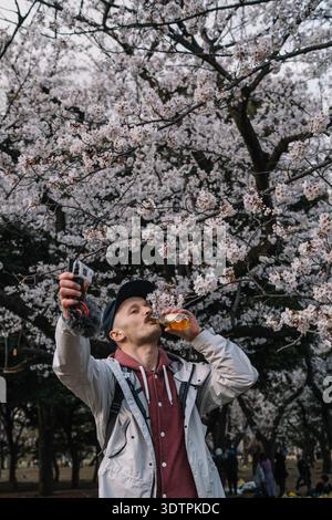 Junger männlicher Blogger, der aus der Flasche trinkt, unter Kirschblüten Stockfoto