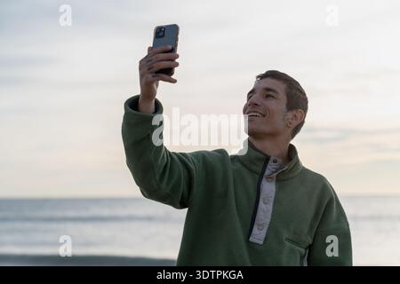 Junger Mann lächelt und nimmt Selfie auf dem Meer Stockfoto