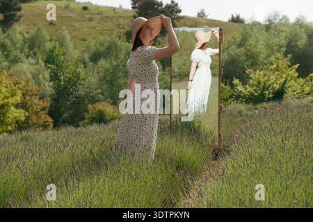 Die junge lächelnde Frau in Blumenkleid und Strohhut steht neben einem großen Spiegel im sonnigen Lavendelfeld und genießt die Natur und Reflexion im Sommer. Stockfoto