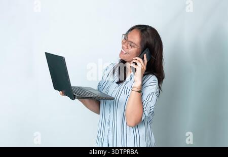 Porträt einer fröhlichen indonesischen Frau mit Brille, die einen Laptop benutzt, während sie mit ihrem Telefon telefoniert. Lächelnder Ausdruck, isoliert auf weißem Hintergrund. Stockfoto