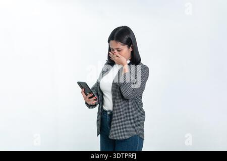 Porträt einer indonesischen Frau in einem karierten Hemd, die mit einem schockierten oder traurigen Ausdruck auf ihr Handy schaut und ihren Mund mit der Hand bedeckt. Isoliert am Stockfoto