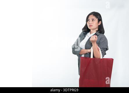Eine indonesische Frau in kariertem Hemd und Jeans, die mit einer roten Tasche steht. Selbstbewusster Ausdruck mit überkreuzten Armen. Isoliert auf weißem Hintergrund. Stockfoto