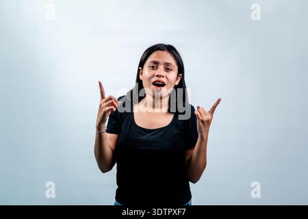 Eine aufgeregte indonesische Frau, die mit offenem Mund in die Kamera schaut und beide Zeigefinger nach oben zeigt. Isoliert auf weißem Studiohintergrund. Stockfoto