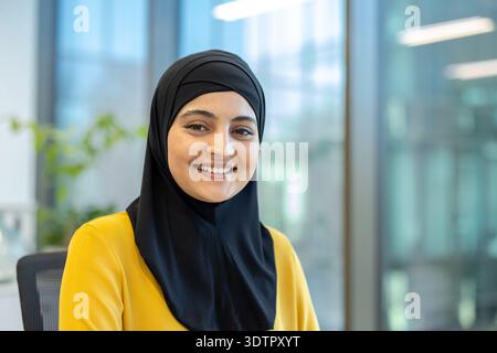 Muslimische Frau mit Hijab und gelbem Oberteil, die einen selbstbewussten und freundlichen Ausdruck zeigt, während sie in einer modernen Büroumgebung sitzt und für Vielfalt und Professionalität steht Stockfoto