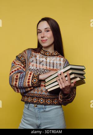Eine selbstbewusste junge Frau steht stolz mit einem Stapel Bücher in den Händen. Ihr farbenfroher Pullover sorgt für Wärme auf dem hellgelben Hintergrund und schafft so etwas Stockfoto