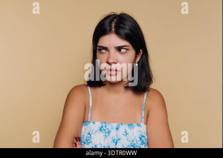 Eine junge Frau mit dunklem Haar steht vor einem weichen beigen Hintergrund, ihre Arme überkreuzt und Ausdruck offenbart Verwirrung und Kontemplation. Der Moment Stockfoto