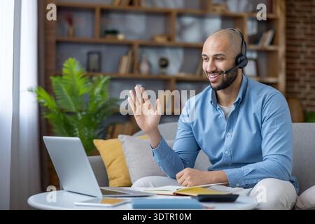 Mann lächelt und winkt mit der Hand, während er eine virtuelle Videokonferenz auf einem Laptop abhält, von einem komfortablen Home Office aus arbeitet, online kommuniziert Stockfoto