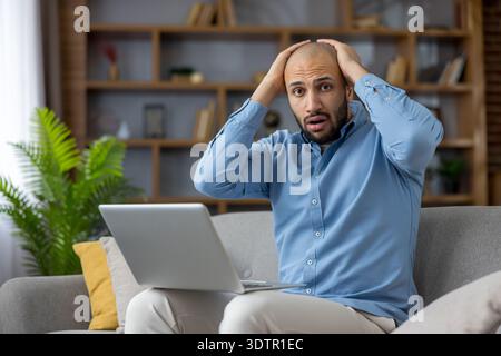 Junger Mann, der verzweifelt den Kopf hält, sich gestresst und schockiert fühlt, nachdem er schlechte Nachrichten auf dem Laptop-Bildschirm gesehen hat und Frustration und Sorgen erlebt, während er auf dem Sofa sitzt Stockfoto