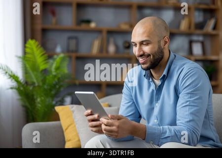 Glücklicher junger Mann mit Bart, der sich auf einem modernen Wohnzimmer-Sofa zurücklehnt und lächelt, während er sich mit Freunden per Videoanruf auf einem digitalen Tablet verbindet, um sich online zu unterhalten und zu arbeiten Stockfoto