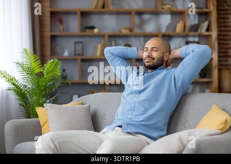 Junger Mann, der ein blaues Hemd und eine leichte Hose trägt, entspannt sich auf einem bequemen grauen Sofa mit Händen hinter dem Kopf und genießt einen friedlichen Moment mit geschlossenen Augen in seinem modernen Wohnzimmer Stockfoto