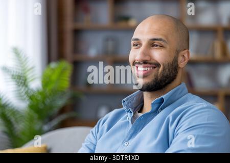 Ein kahler, bärtiger Mann, der im Profil lächelt, entspannt in einem hellen Wohnzimmer sitzt, mit Freude und Zufriedenheit wegblickt und entspanntes Selbstvertrauen und ein Gleichgewicht zwischen Arbeit und Privatleben verkörpert Stockfoto