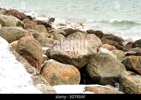 Schneebedeckte Felsen säumen eine zerklüftete Küste, während kalte Wellen vom Meer unter einem bewölkten Winterhimmel hereinströmen. Stockfoto