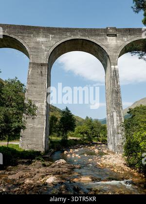 Ein flacher Blick zeigt die massive Betonkonstruktion des Glenfinnan Viadukts, während er sich über ein felsiges Flussbett erstreckt, umgeben von üppigen grünen Bäumen A Stockfoto