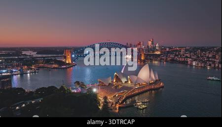 Australien, Sydney: Der Hafen von Sydney bei Sonnenuntergang, das beleuchtete Opernhaus von Sydney und die Harbour Bridge vor dem pulsierenden Himmel. Atemberaubende Aussicht auf die moderne Skyline der Stadt bei Nacht. Drohnenflug Stockfoto